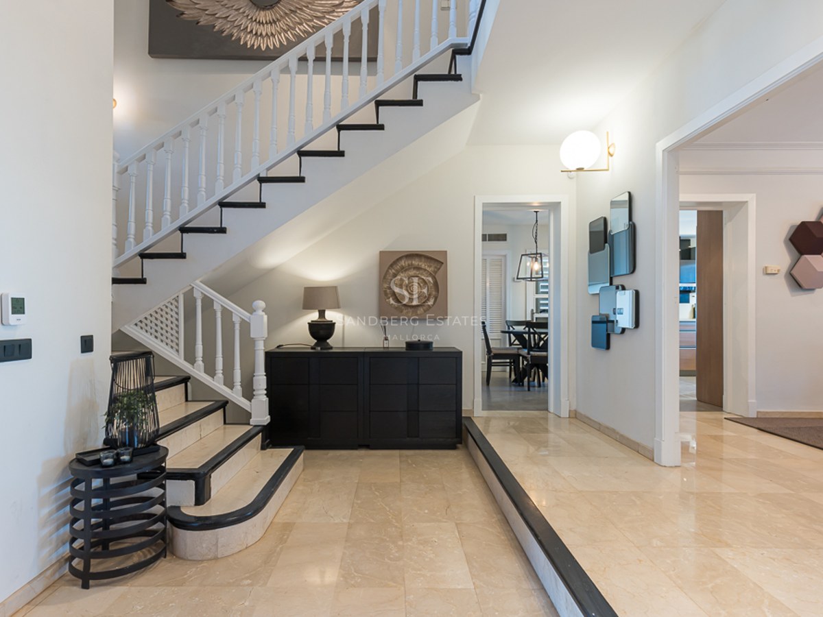 Hall de entrada de lujo con suelos de mármol crema, escalera blanca con detalles negros y decoración contemporánea.