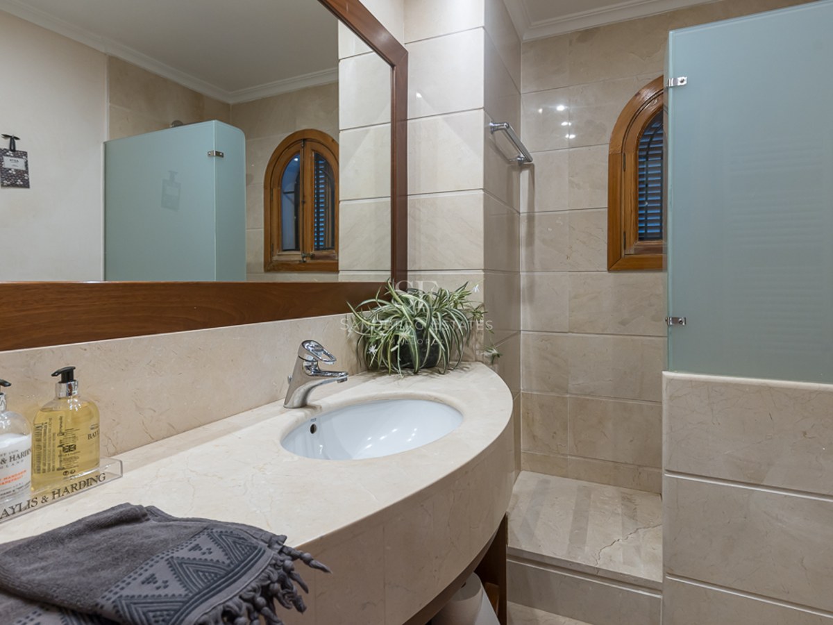 Baño con lavabo de mármol crema, gran espejo con marco de madera y ducha con mampara de cristal ácido.