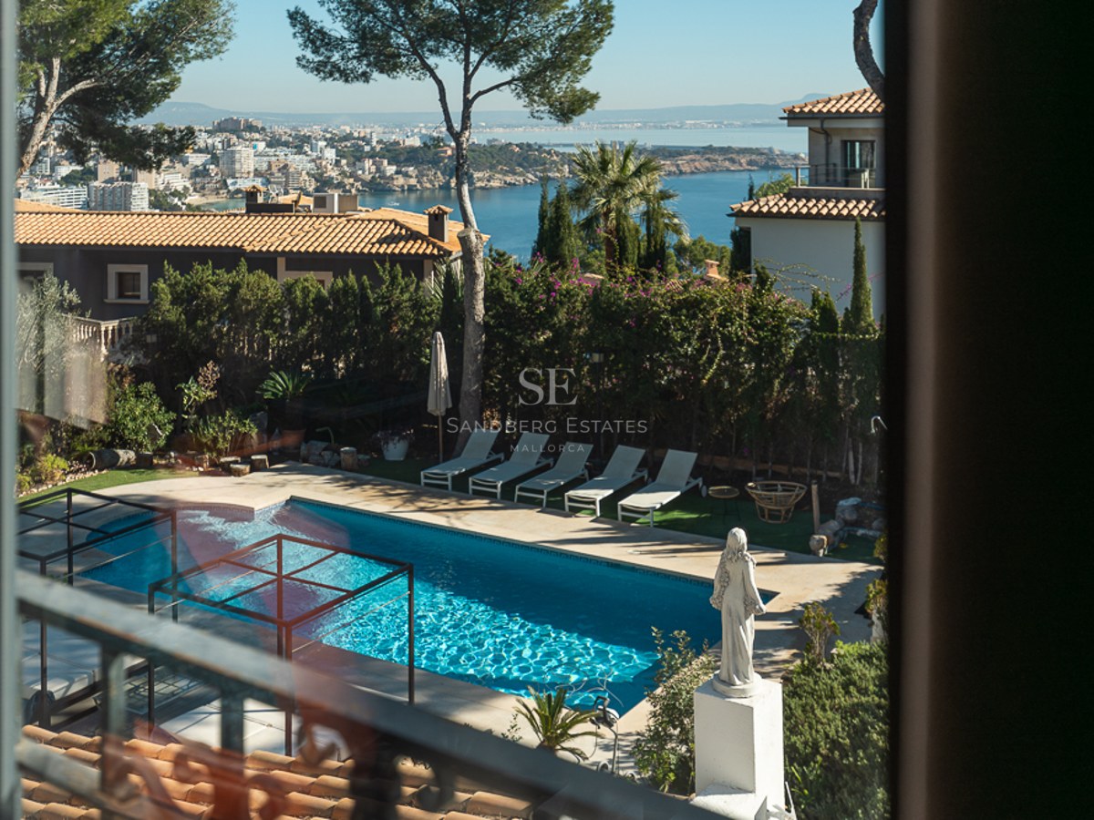 Vista desde una planta superior de una piscina turquesa, tumbonas y la bahía mediterránea al fondo bajo un cielo despejado.