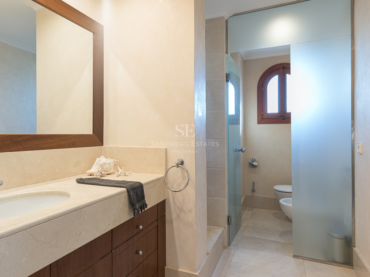 Baño moderno con encimera de mármol crema, muebles de madera oscura y panel de cristal translúcido.