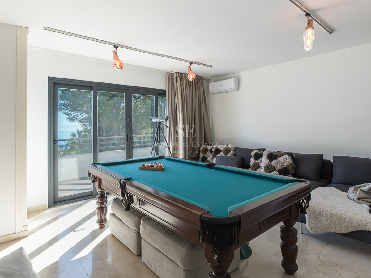Sala de juegos con mesa de billar de madera, sofás grises y puertas correderas de cristal con vistas al exterior.