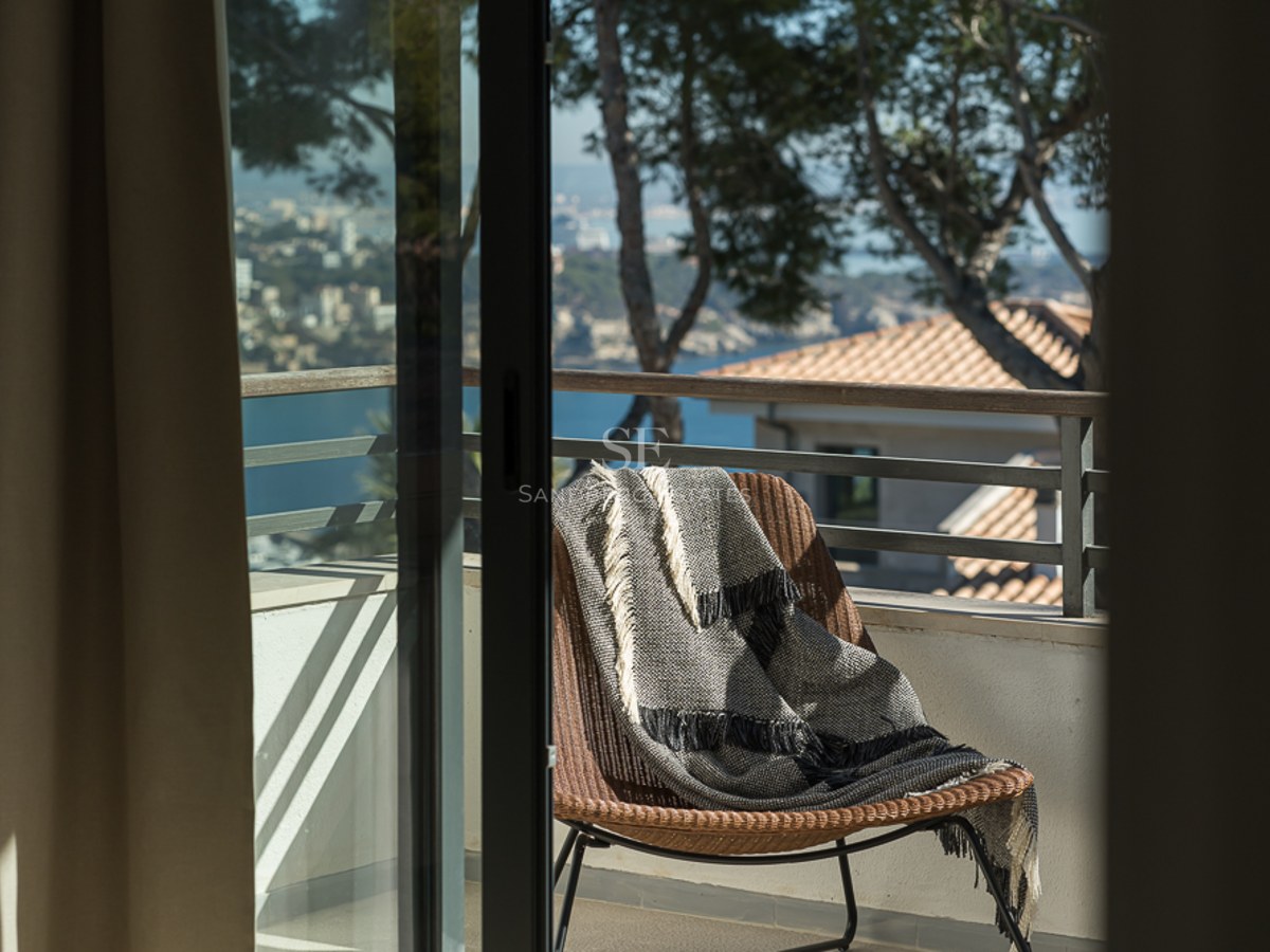 Vista desde una puerta abierta a un balcón con silla de mimbre, manta y vistas a la costa mediterránea al fondo.