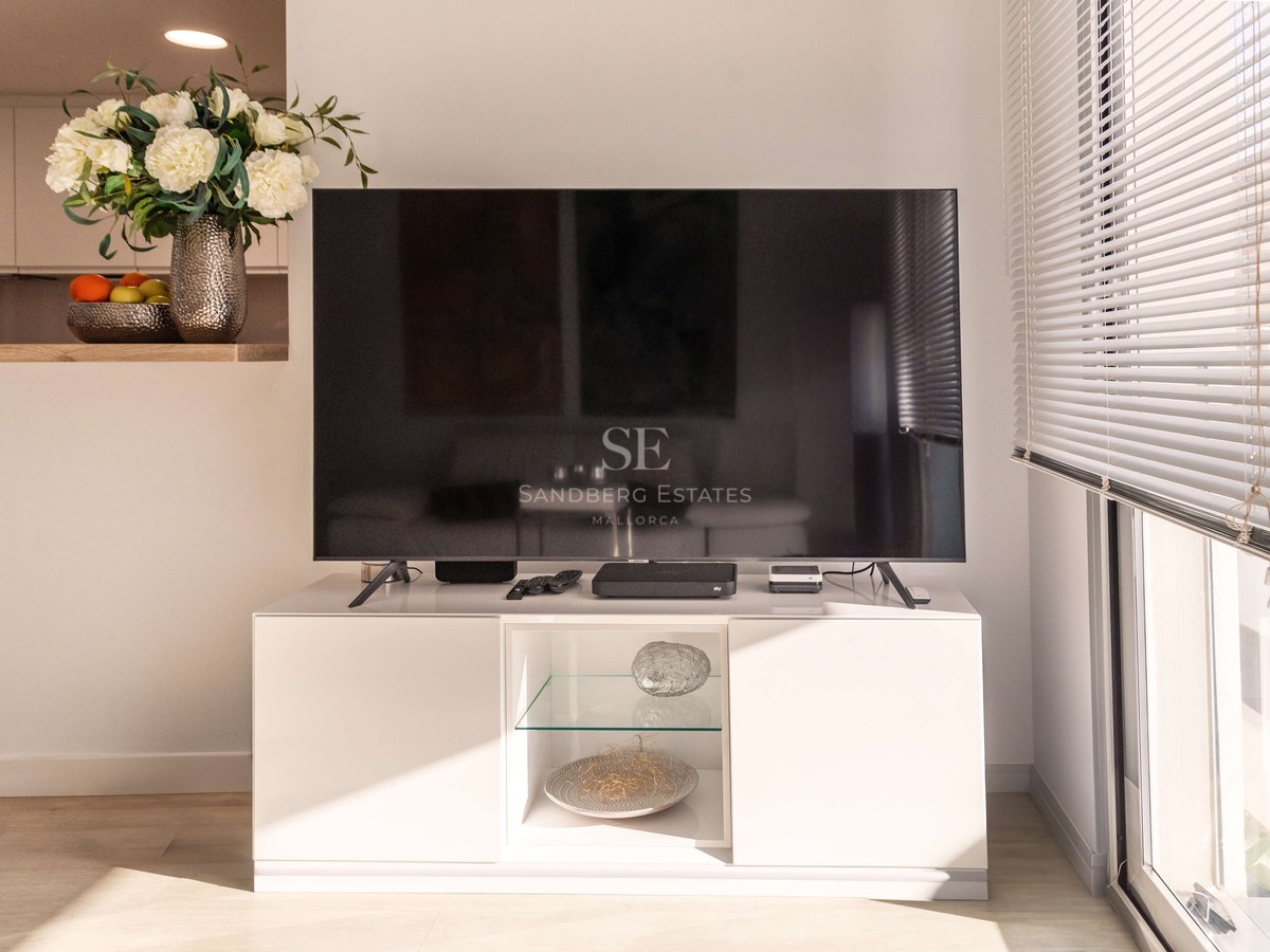 Grand téléviseur sur un buffet blanc minimaliste à côté d'une fenêtre lumineuse avec stores et décor floral.