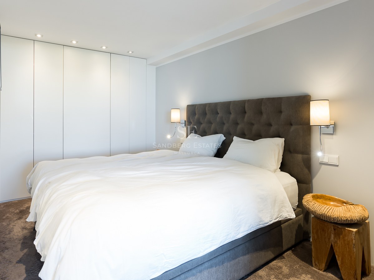 Chambre contemporaine avec lit blanc, tête de lit grise capitonnée, placards blancs et table de chevet en bois brut.