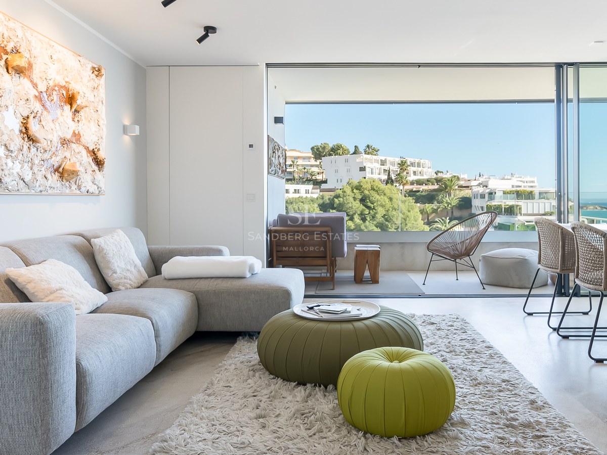 Salon moderne et lumineux avec canapé gris et poufs verts, doté de baies vitrées offrant une vue sur la mer.