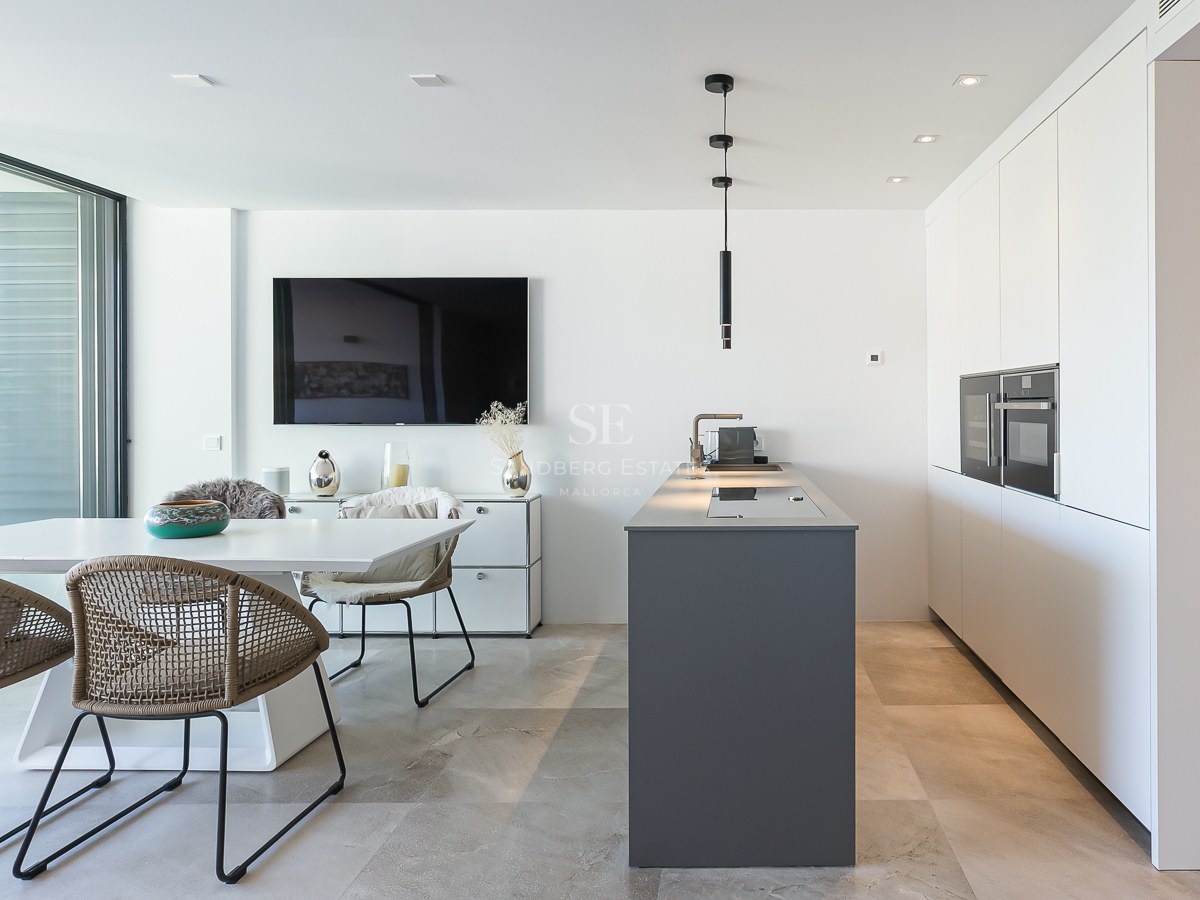 Cuisine blanche moderne avec îlot gris, table à manger et grandes baies vitrées donnant sur la mer.