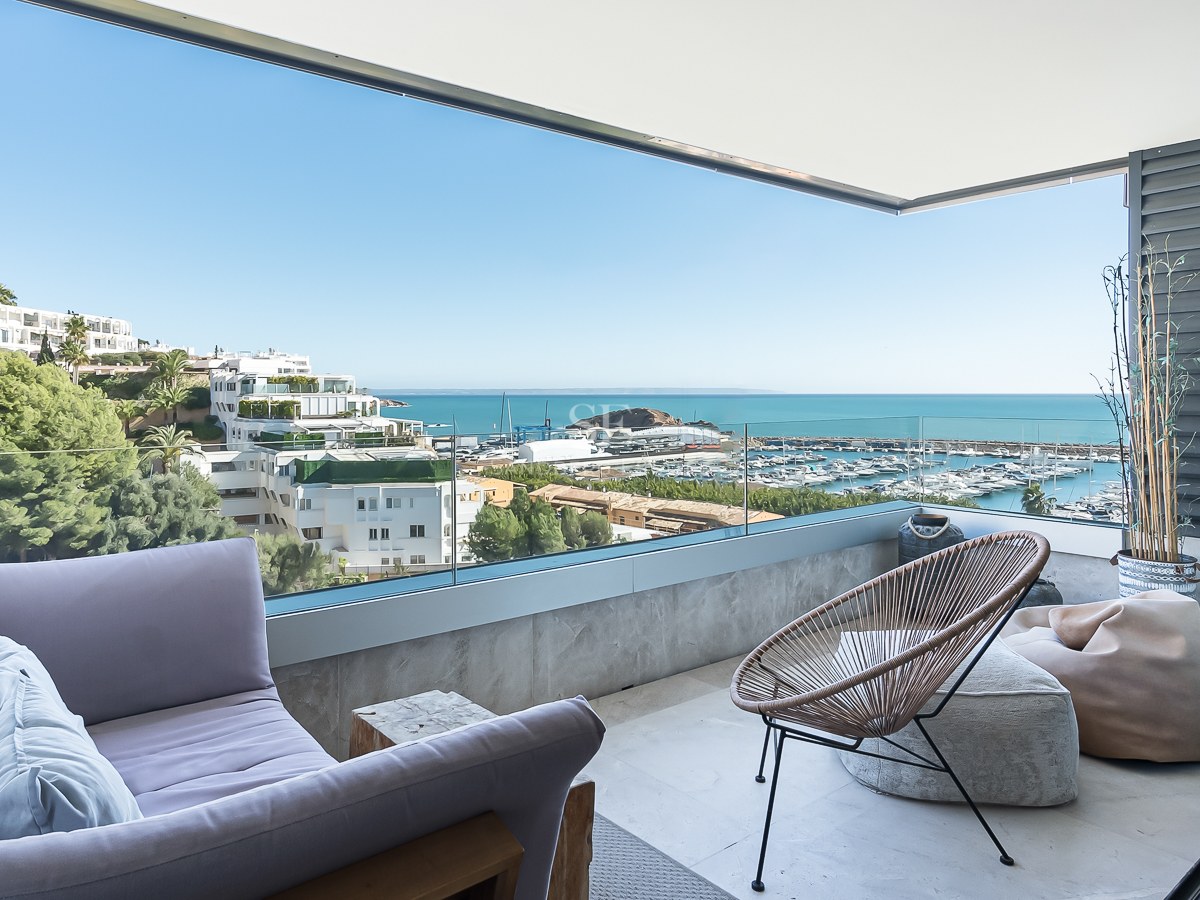 Terrasse couverte avec canapé lavande, fauteuil en osier et garde-corps en verre surplombant la marina et la mer.