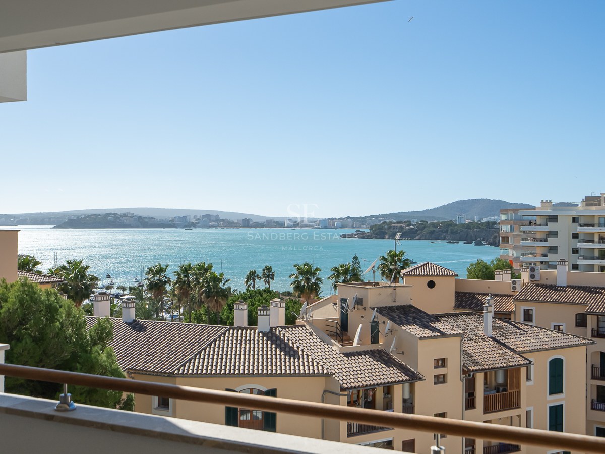 Vue d'un balcon surplombant des toits méditerranéens, des palmiers et une vaste baie turquoise sous un ciel bleu clair.