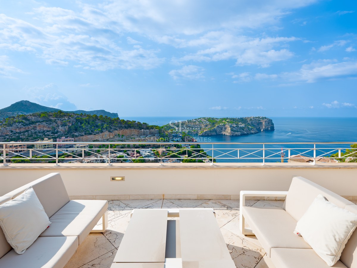 Elevated terrace with white outdoor sofas and a metal railing overlooking a blue bay and rocky Mediterranean coastline.