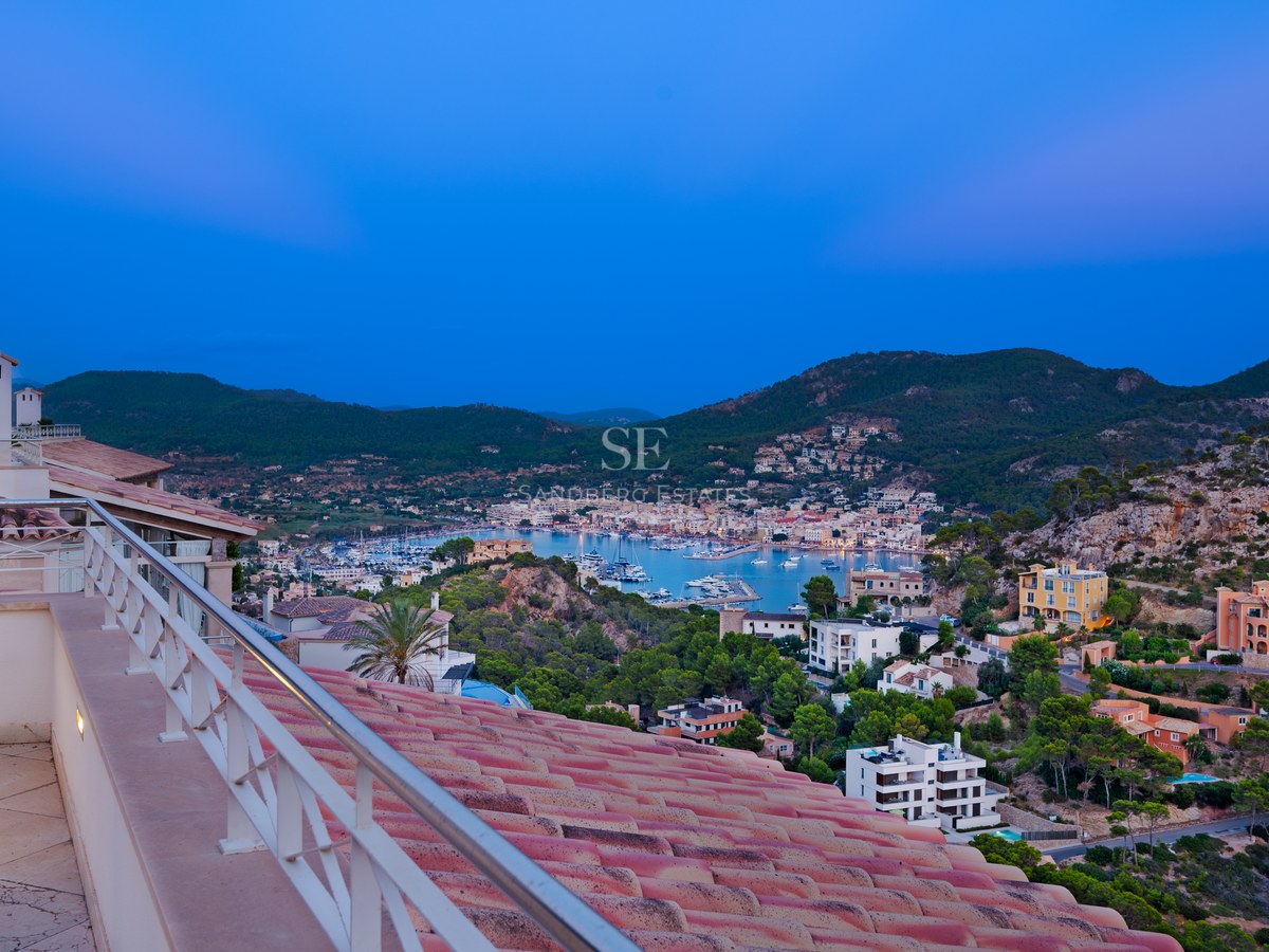 Elevated evening view of a Mediterranean harbor with boats, terracotta roofs in foreground, and mountains beyond.