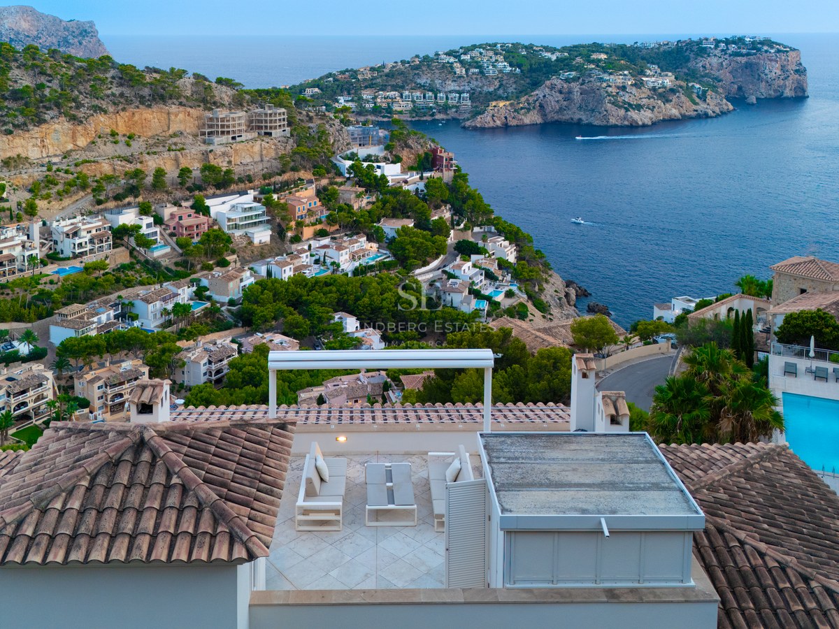 Elevated view of a white modern rooftop lounge with sofas overlooking a Mediterranean bay and coastal hills.