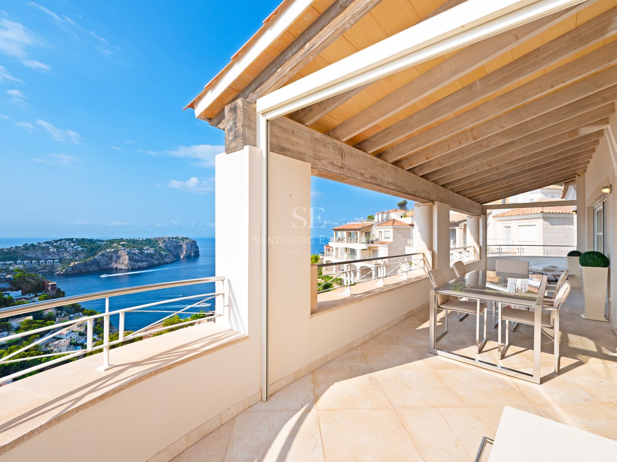 A sunny covered terrace with wooden beams and a dining area overlooking a blue sea and rocky coastline.