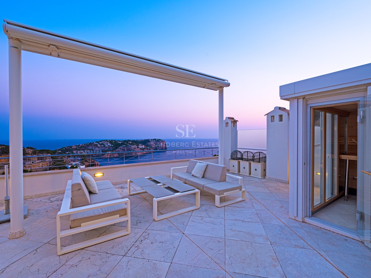 Modern rooftop terrace with white outdoor sofas overlooking a coastline at sunset.