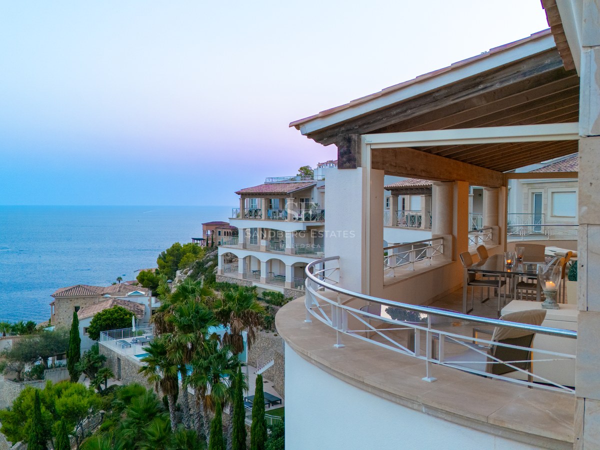 Curved stone balcony with stainless steel railings overlooking Mediterranean villas and the sea at sunset.