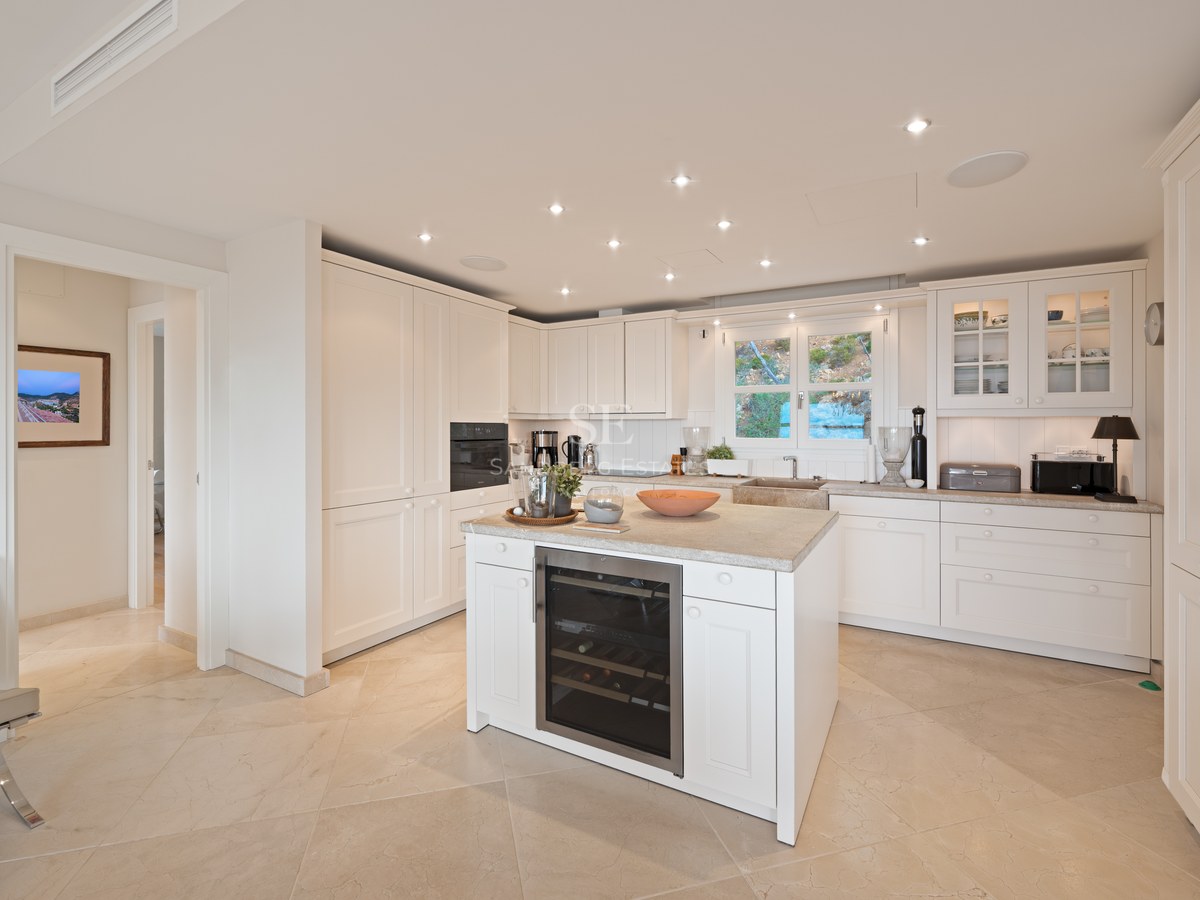 Bright white kitchen featuring a central island with integrated wine fridge, stone flooring, and recessed LED lighting.