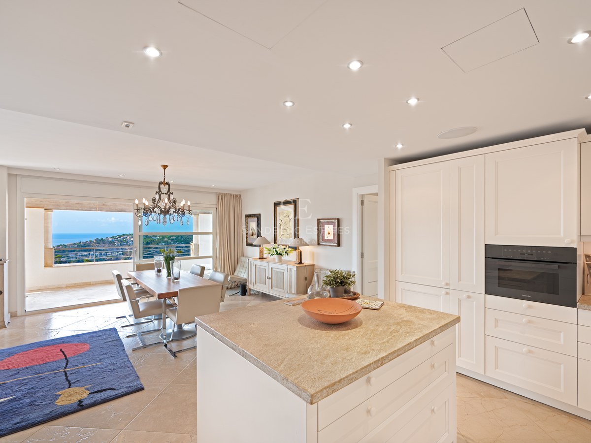Modern white kitchen island and dining table in a bright room overlooking the sea through glass sliding doors.