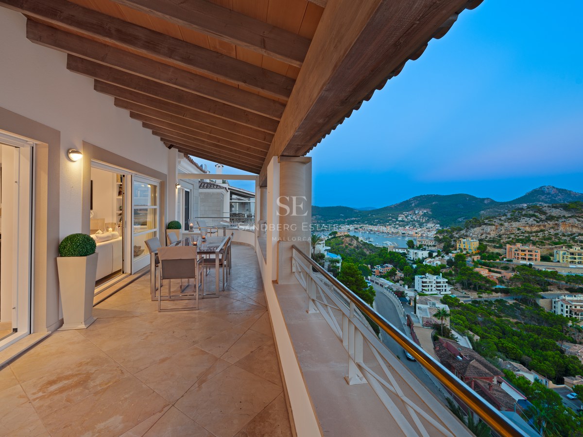 A spacious covered terrace with wooden ceiling beams, stone flooring, and a dining set overlooking a coastal town at dusk.
