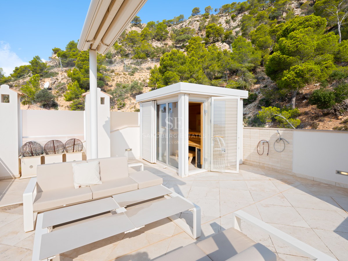 A sunny white stone terrace featuring an outdoor sauna cabin, lounge seating, and a backdrop of pine trees and rocky hills.