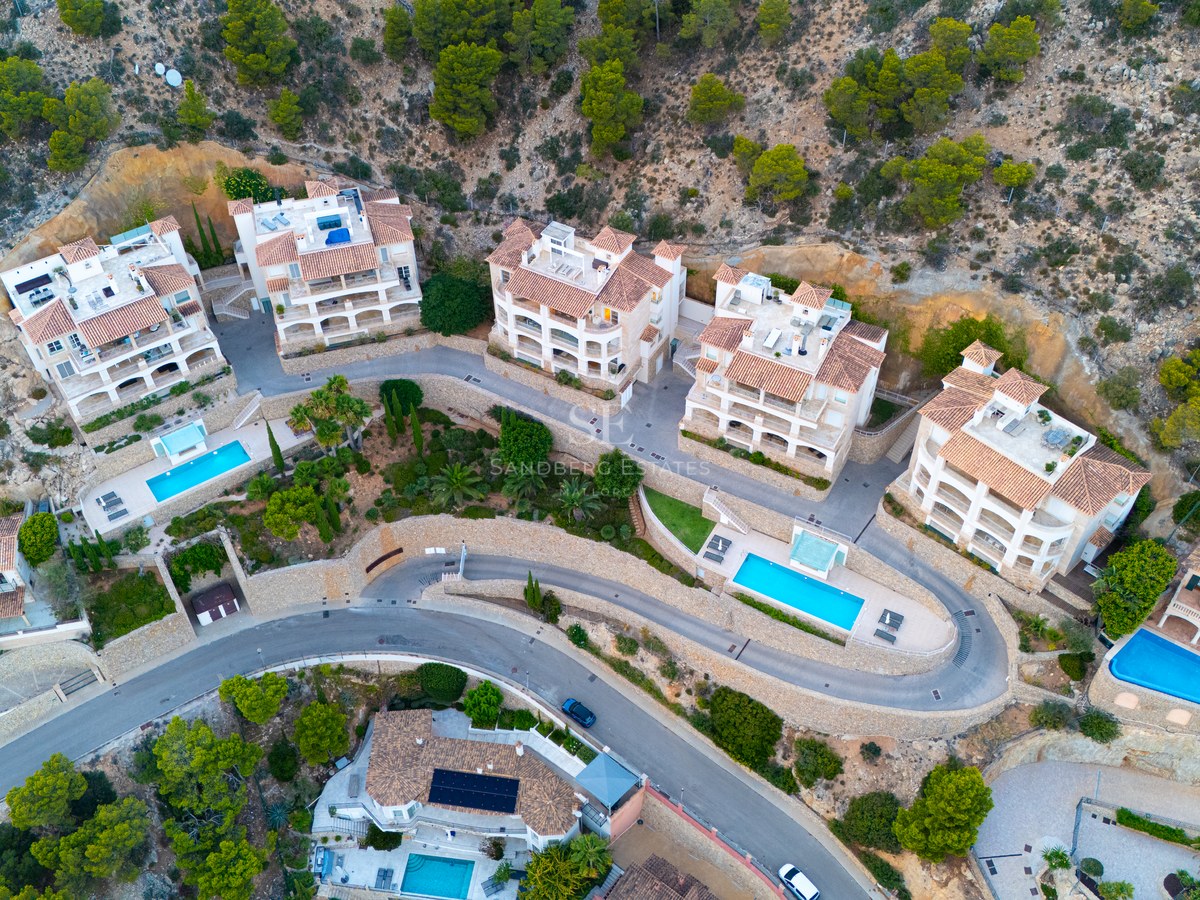 Overhead drone shot of luxury apartments with terracotta roofs and turquoise pools nestled in a green hillside.