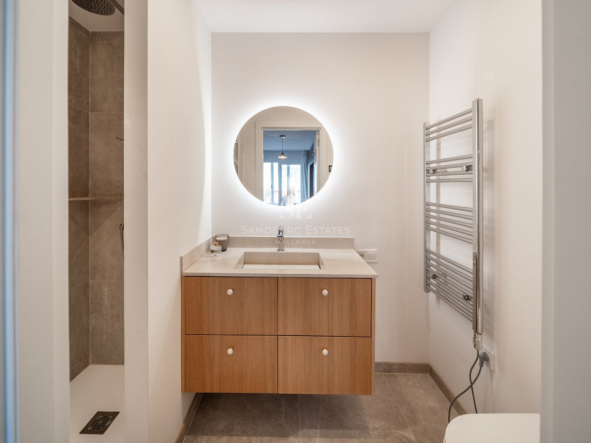 Salle de bain minimaliste avec miroir circulaire rétroéclairé, meuble en chêne, douche à l'italienne et sèche-serviettes.