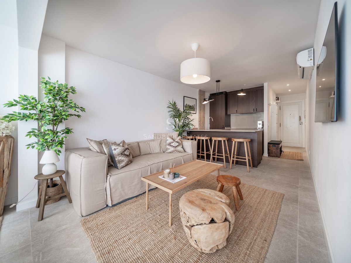 Salon lumineux à aire ouverte avec canapé beige, table basse en bois, tapis en jute et comptoir de cuisine.