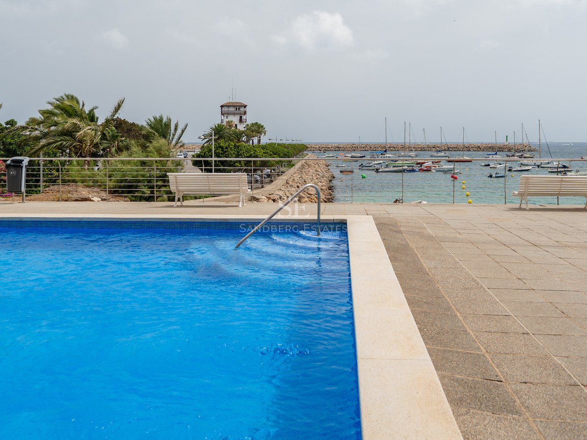 Piscine bleue avec rampe en inox donnant sur un port de plaisance avec bateaux et promenade en pierre.