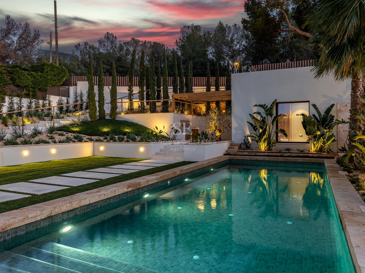 A lit swimming pool with turquoise water at dusk, surrounded by stone paving, green lawns, and tropical plants.