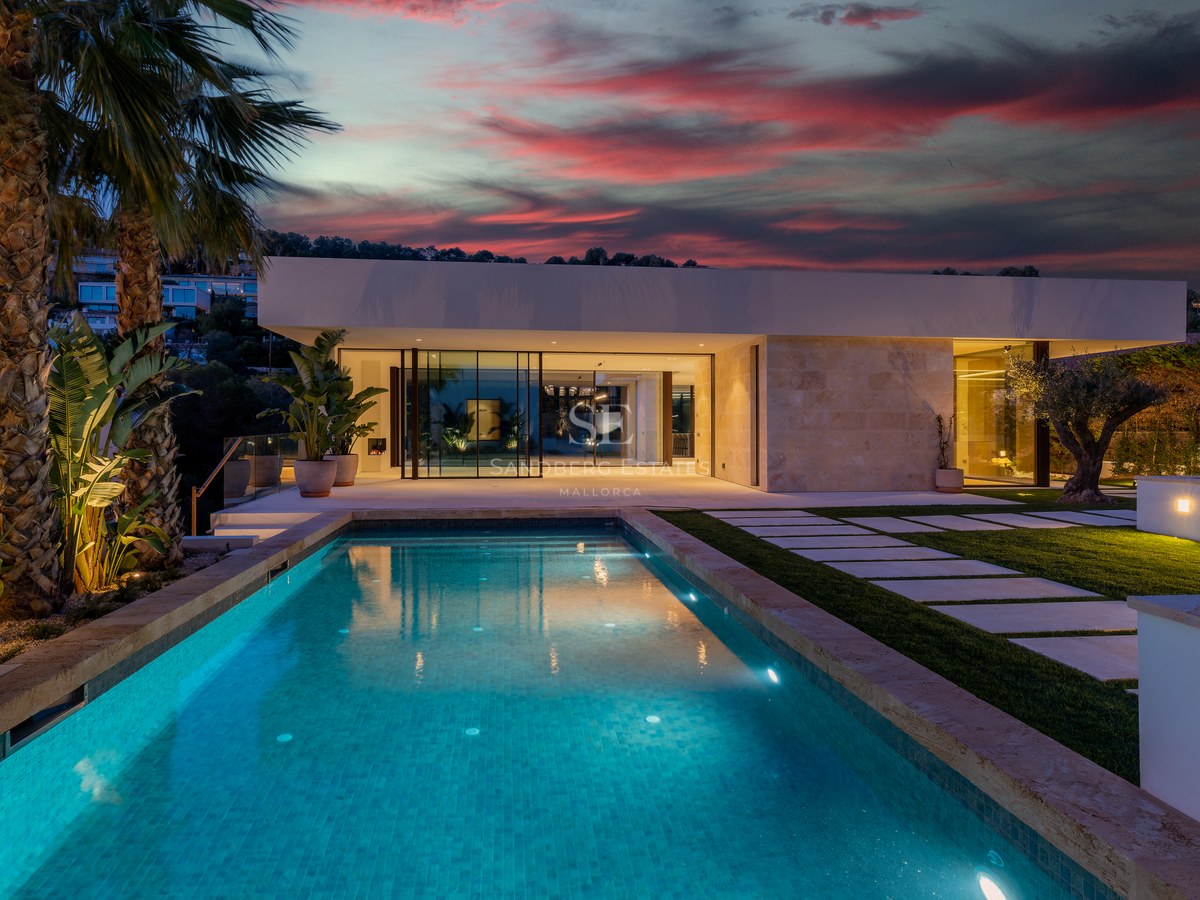 Illuminated swimming pool in front of a modern natural stone villa under a dramatic red sunset sky with palm trees.