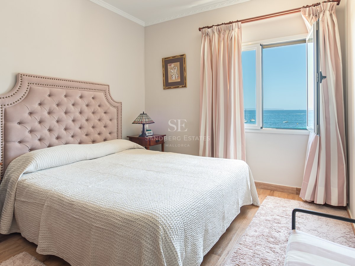 Chambre lumineuse avec tête de lit rose, meubles en bois et fenêtre ouverte donnant sur la mer Méditerranée.