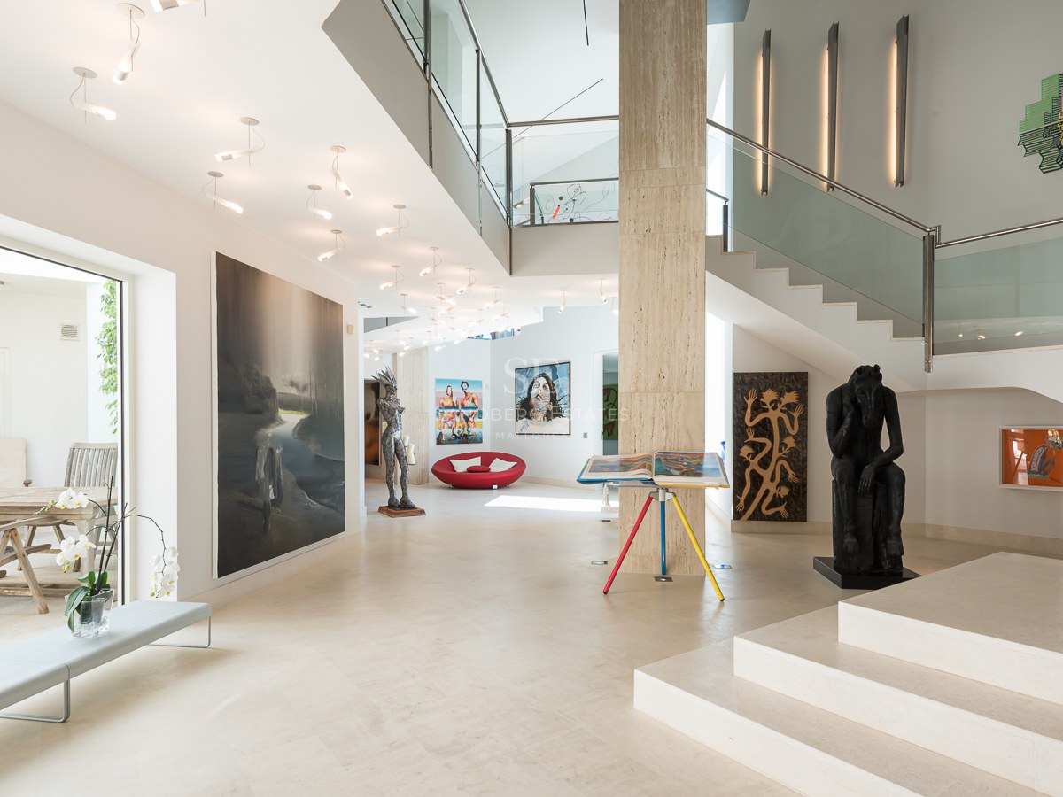 Spacious modern entrance hall with cream travertine floors, glass staircase, and large-scale contemporary artworks.