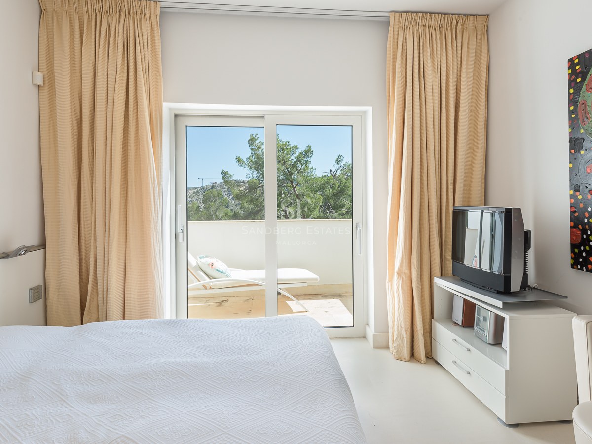 A bright bedroom featuring a white bed, beige curtains, and sliding glass doors leading to a terrace with a lounger.