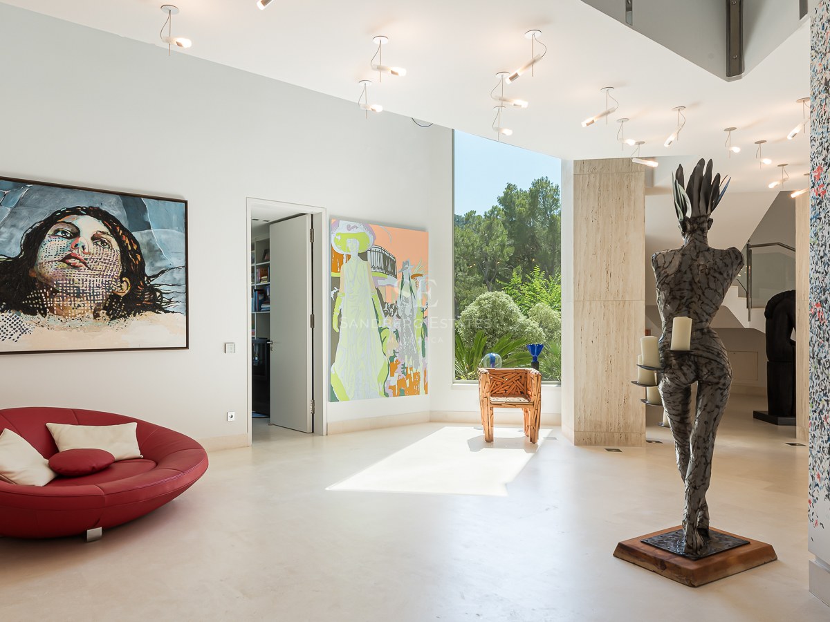 Modern white hallway with large pop-art paintings, a red circular lounge chair, and a floor-to-ceiling window.