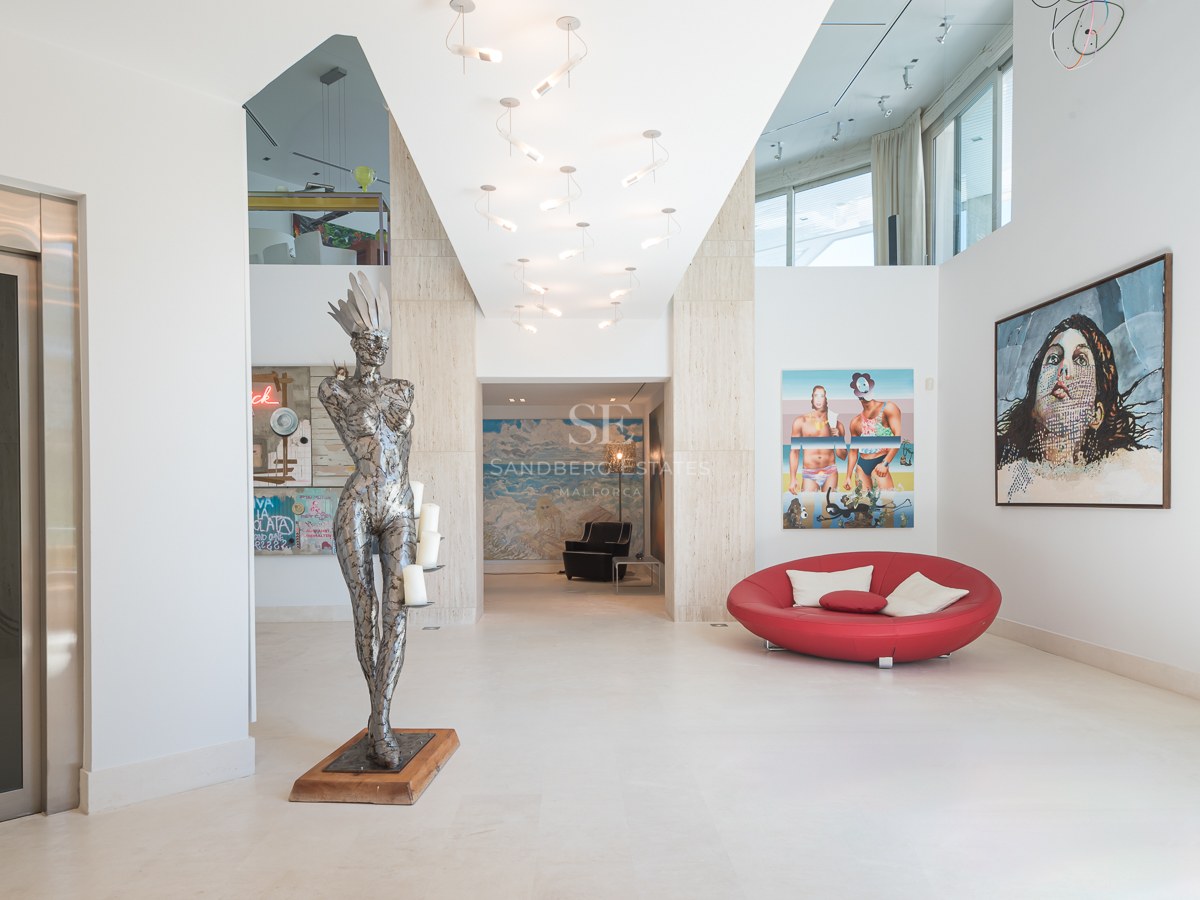 Modern white entrance hall with a private elevator, metal sculpture, red sofa, and large contemporary art pieces.