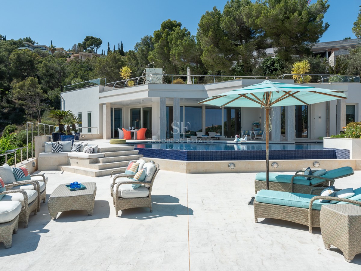 Outdoor terrace with turquoise sunbeds, wicker furniture, and a dark blue mosaic pool overlooking green hills.
