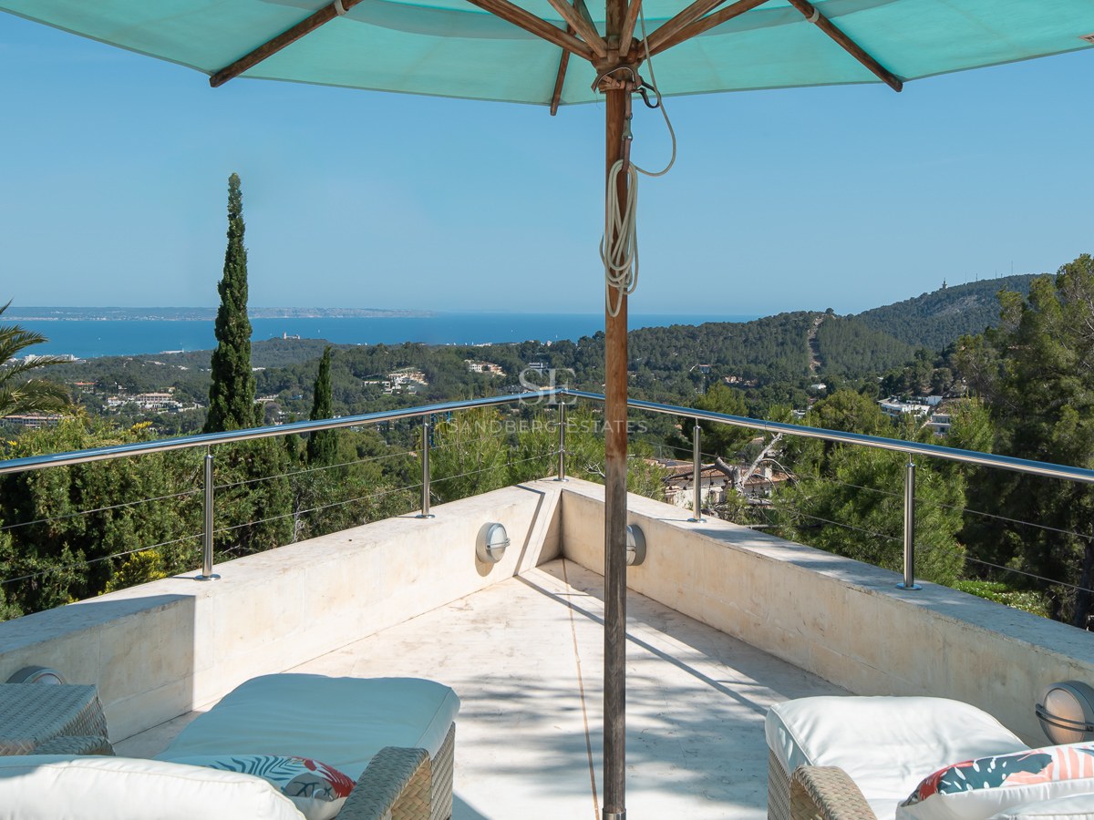 Elevated natural stone terrace with wicker loungers and turquoise parasol overlooking lush pine trees and the sea.
