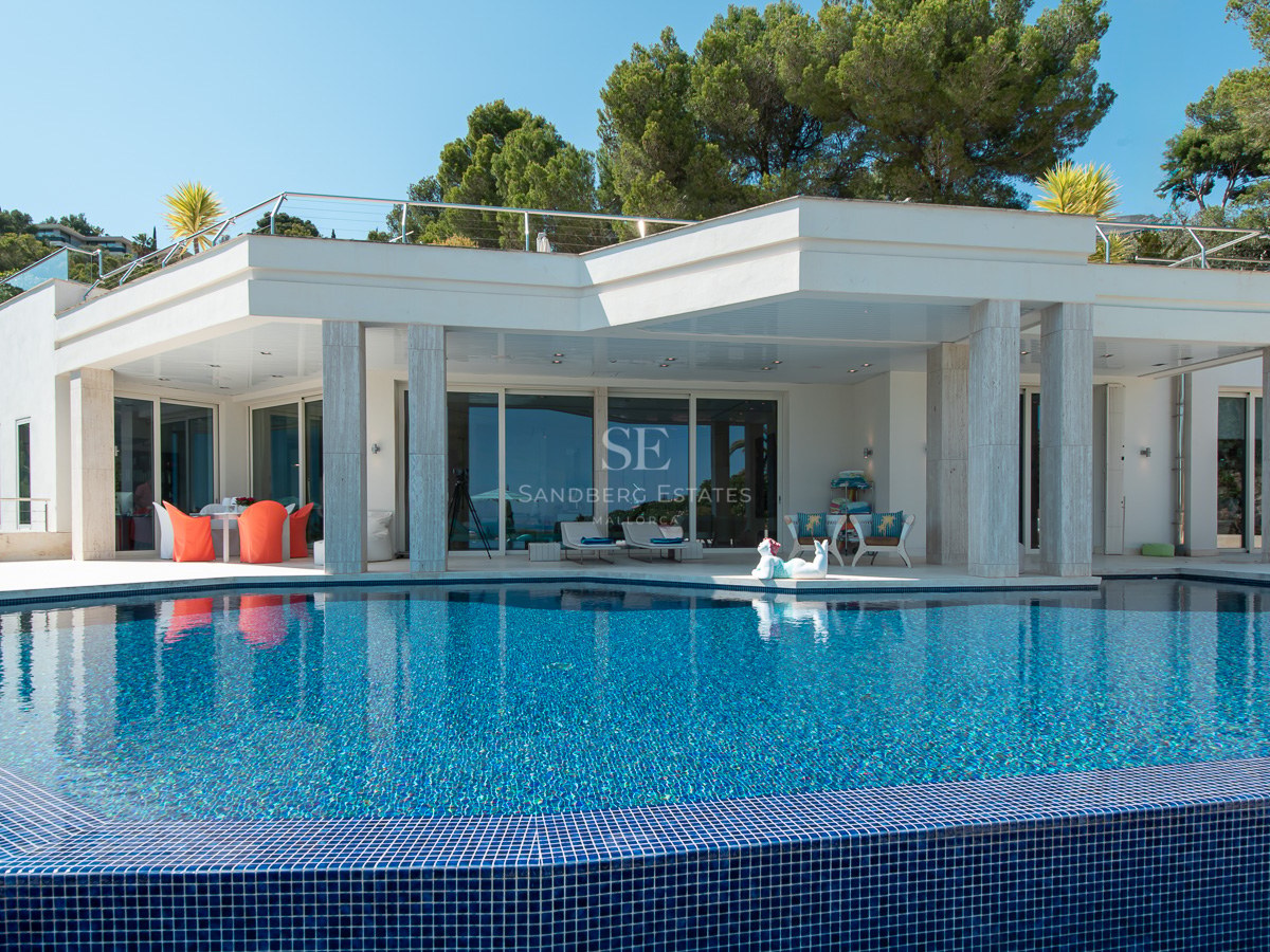 Modern white villa with a blue tiled swimming pool and a covered terrace with marble pillars surrounded by pine trees.