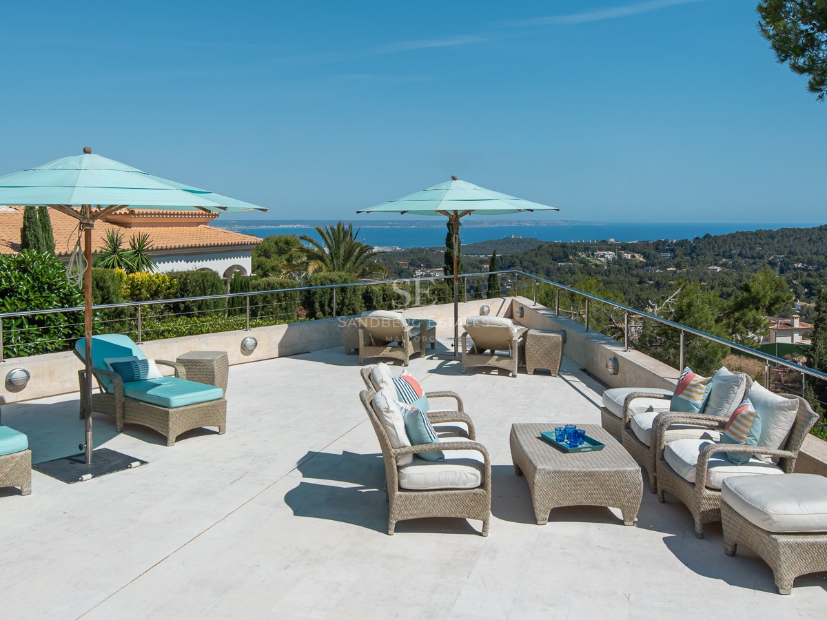 Elevated stone terrace with wicker lounge furniture and teal umbrellas overlooking a pine forest and the sea.