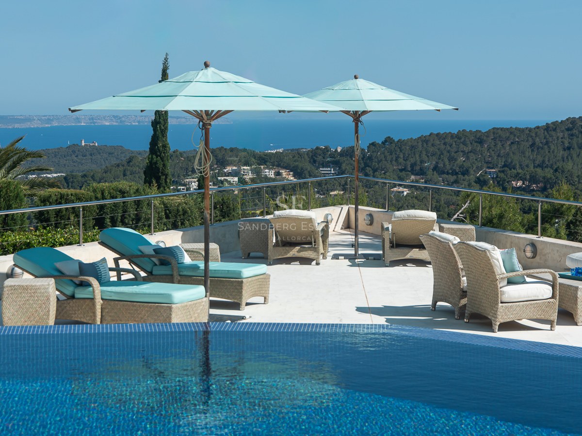 Infinity pool edge overlooking a large stone terrace with wicker furniture and light blue parasols facing the sea.