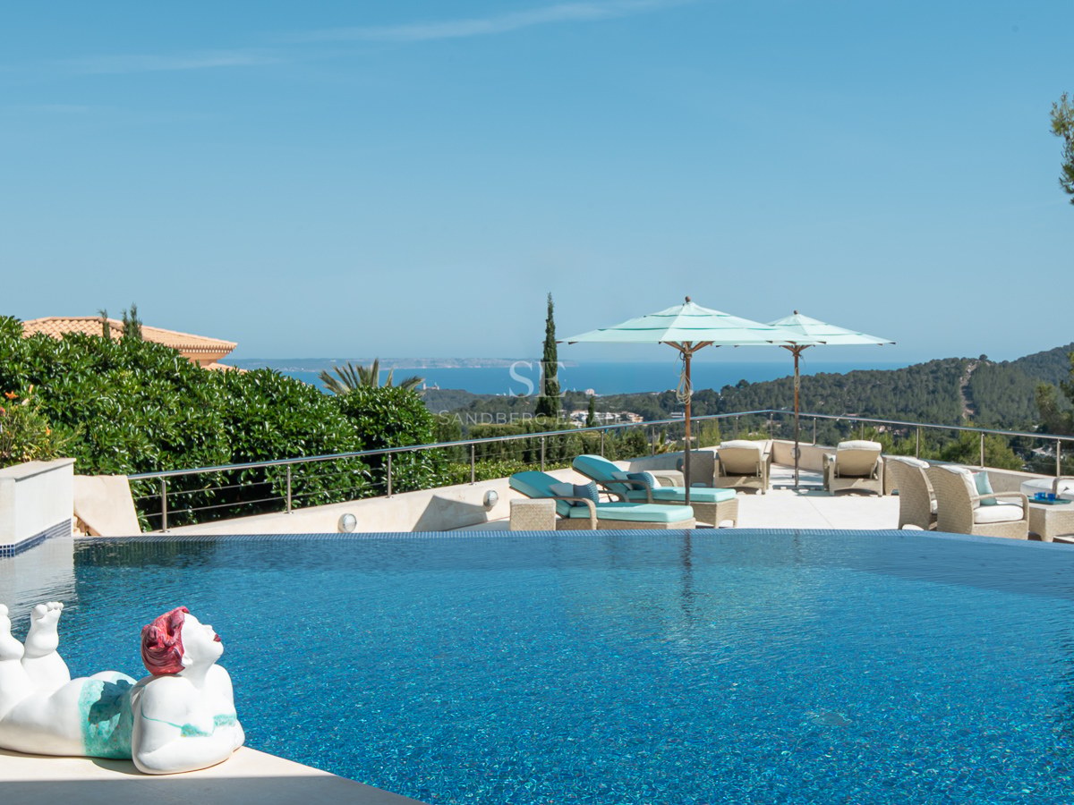 An infinity pool with blue mosaic tiles overlooking the Mediterranean coastline, featuring lounge chairs and a sculpture.