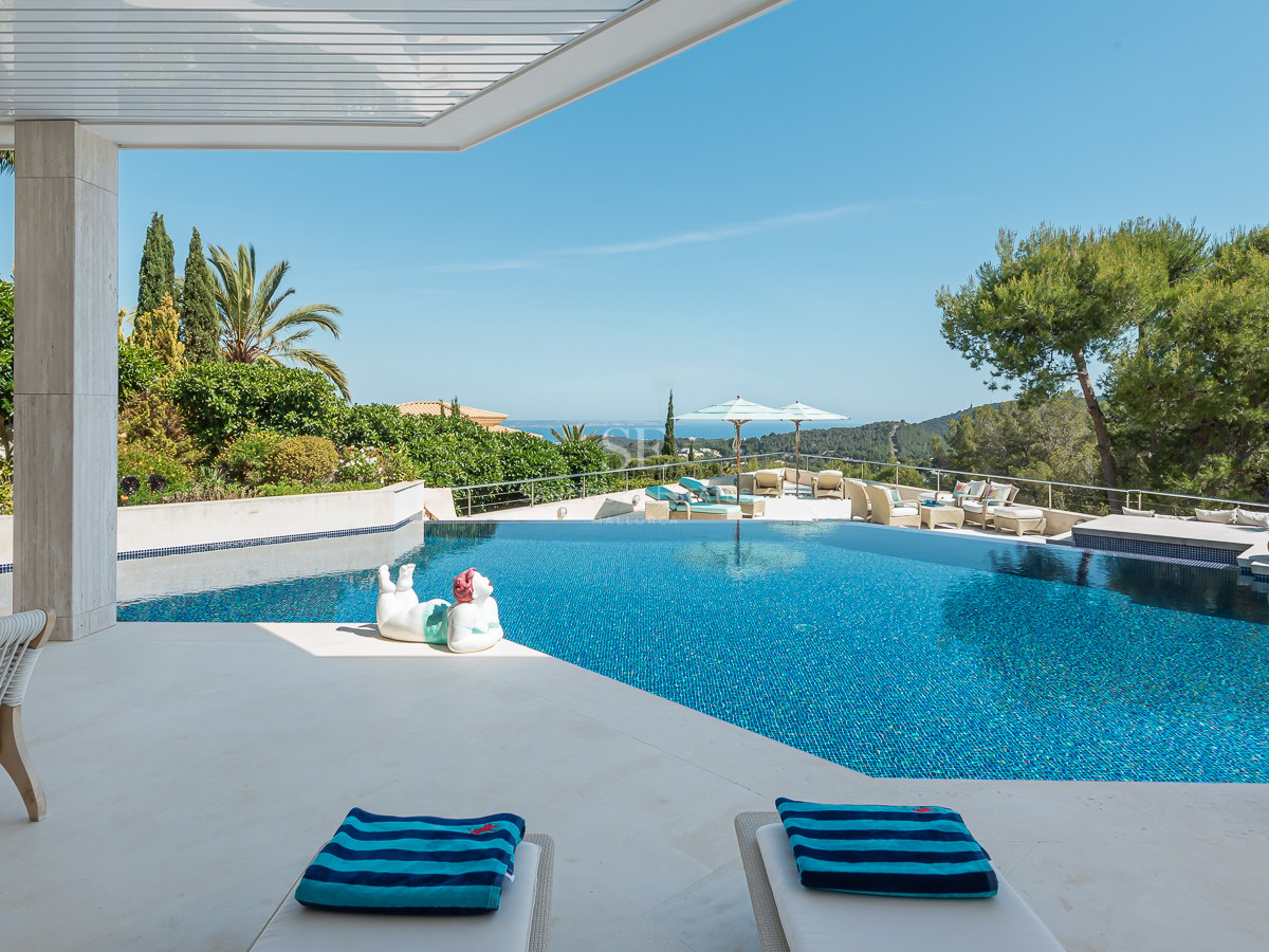 Luxury stone terrace featuring an azure infinity pool, lounge chairs, and panoramic sea views under a clear sky.