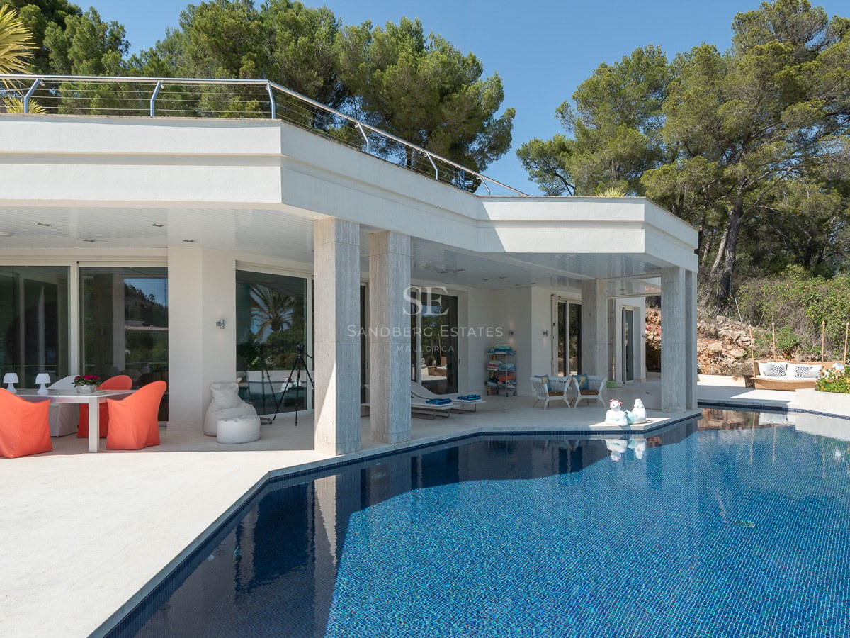 Exterior view of a white modern villa featuring a large blue swimming pool and shaded outdoor dining area.