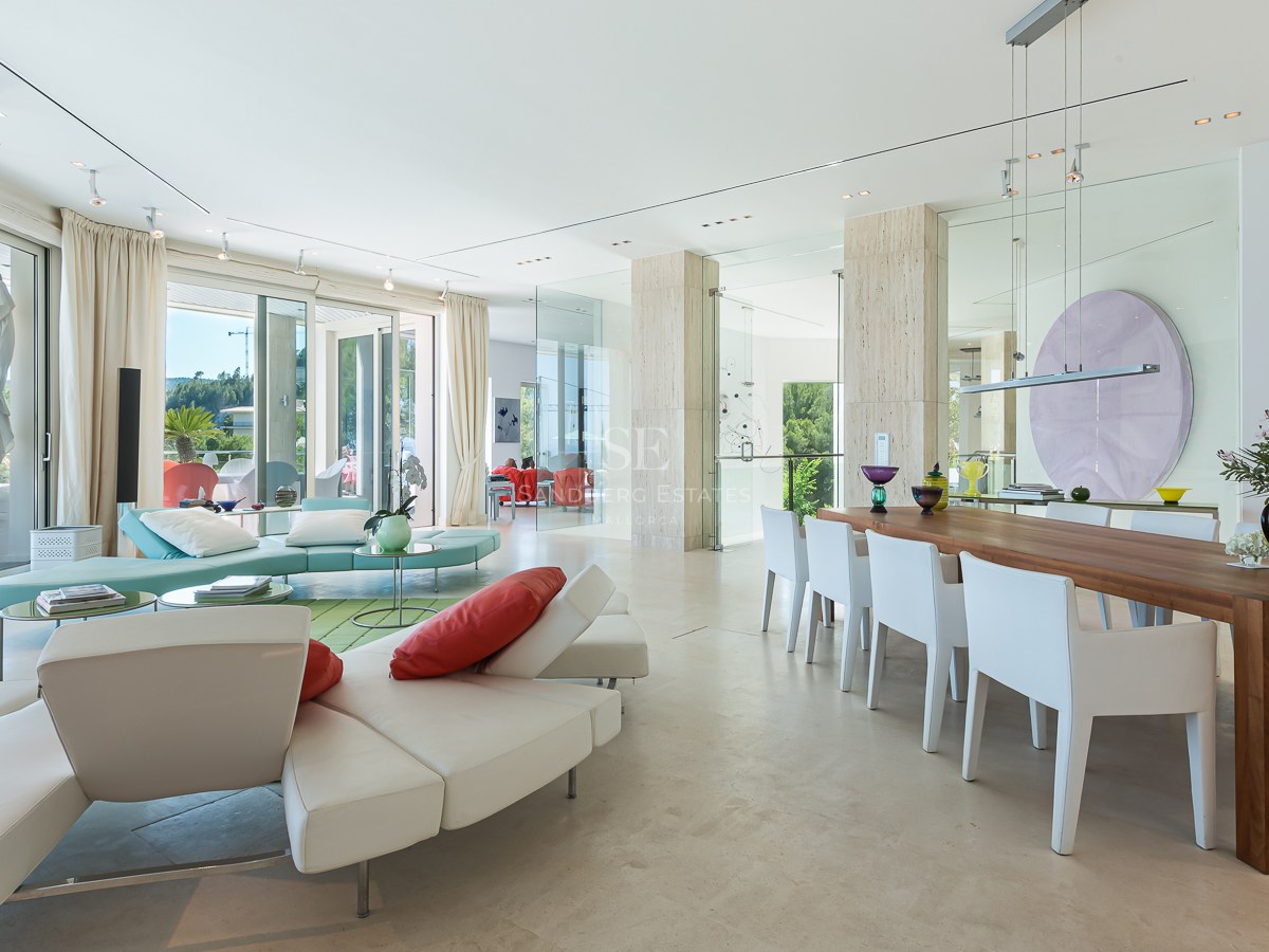 Spacious modern living room with cream stone floors, large glass windows, white leather sofas and a wood dining table.