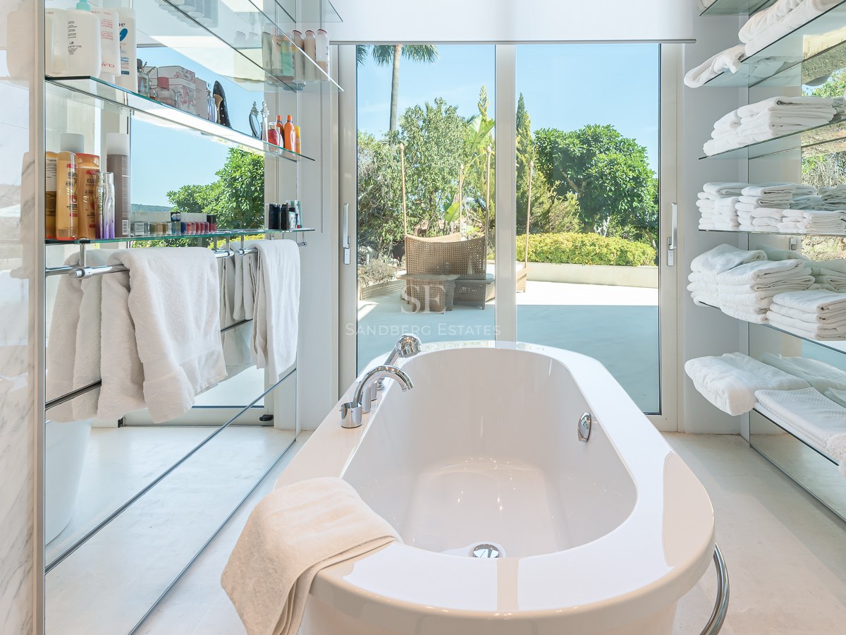 White freestanding bathtub centered between glass shelves, looking out through glass doors to a sunny garden.