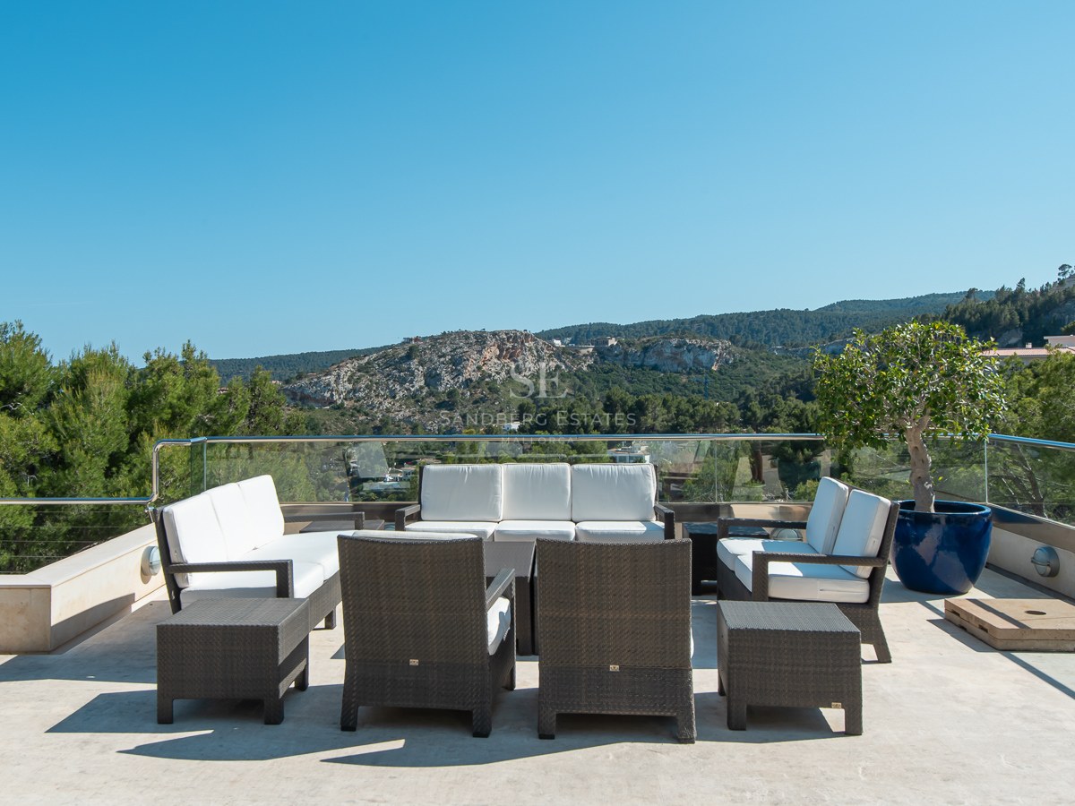 Spacious stone terrace with wicker seating area overlooking a lush mountain landscape under a clear blue sky.