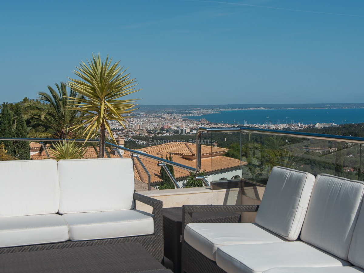 Luxury terrace with white sofas and glass railing overlooking a coastal city and the sea under a clear blue sky.