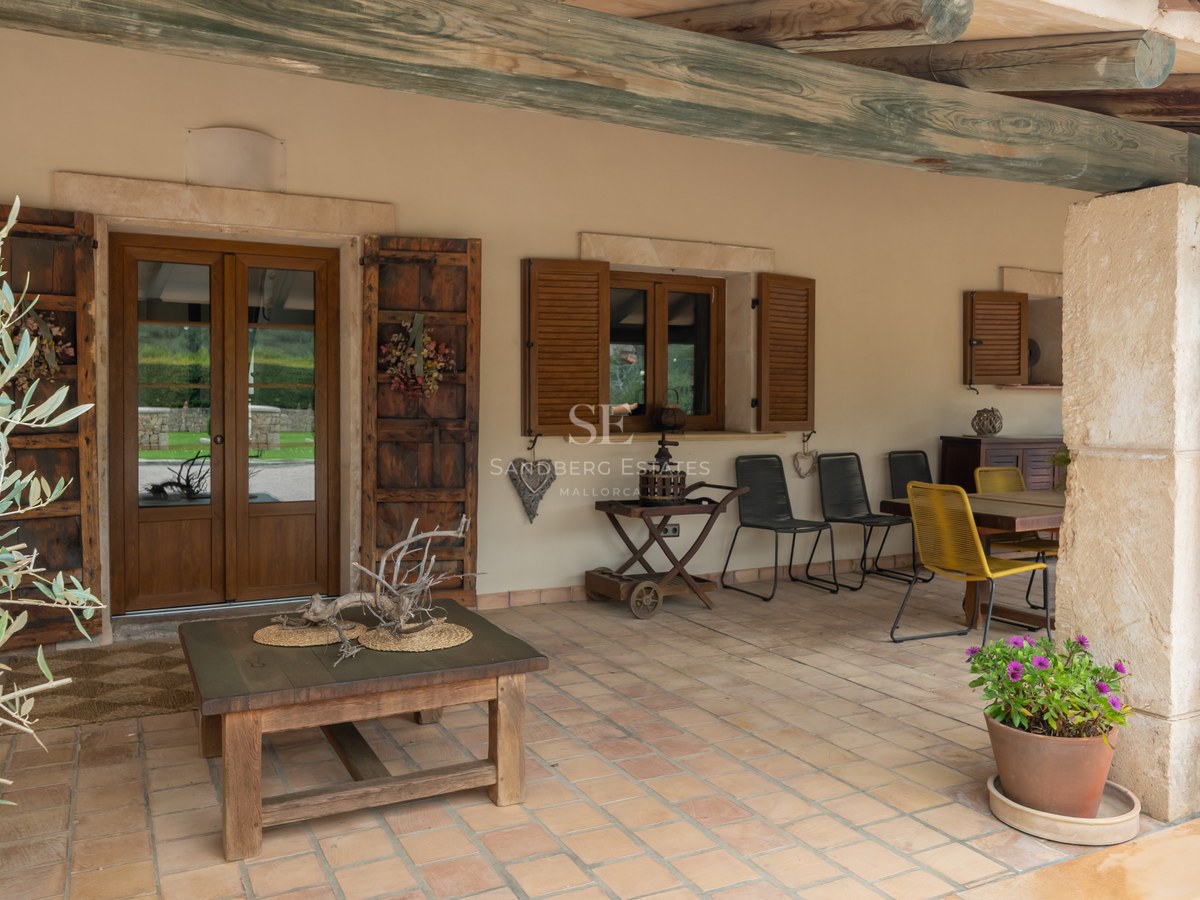 Porche couvert avec carrelage en terre cuite, poutres en bois rustiques, coin repas et murs en pierre claire.
