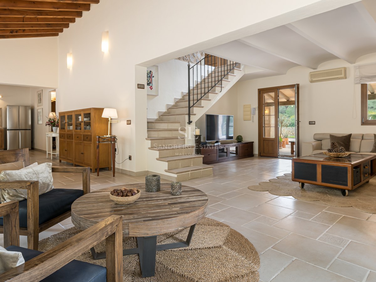 Grand salon avec sol en pierre naturelle, poutres en bois au plafond, escalier en pierre et mobilier classique.
