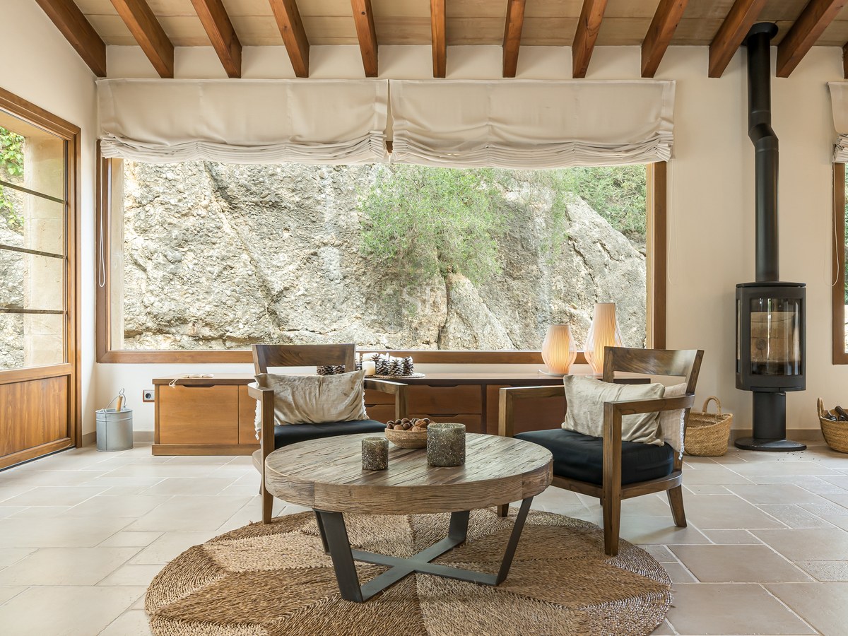 Salon lumineux avec poutres en bois, poêle à bois moderne et vue sur une paroi rocheuse naturelle.