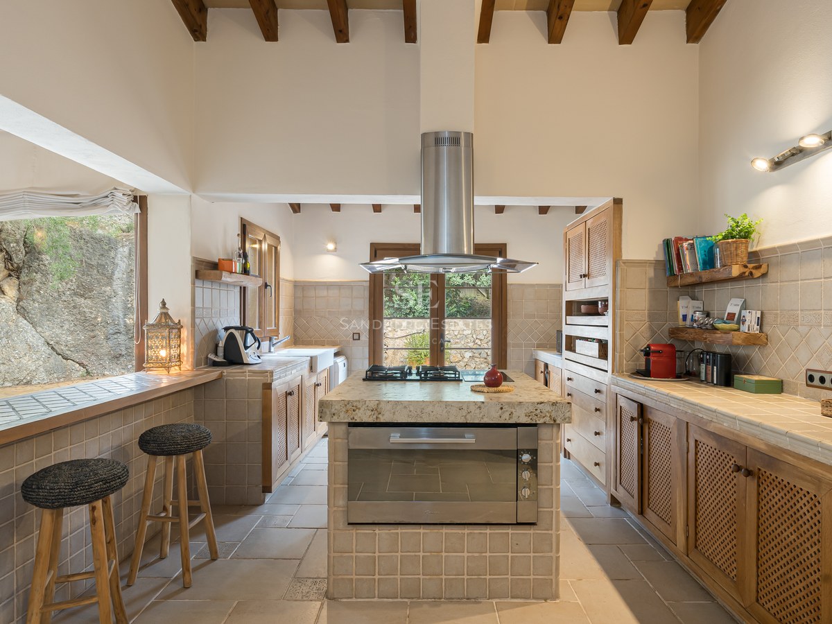 Cuisine rustique avec poutres apparentes, îlot central, plans de travail carrelés et taburets près d'une fenêtre.