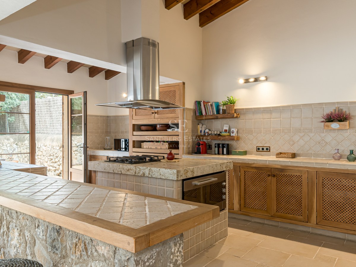 Grande cuisine avec poutres en bois, comptoir en pierre naturelle et îlot carrelé avec plaques de cuisson.